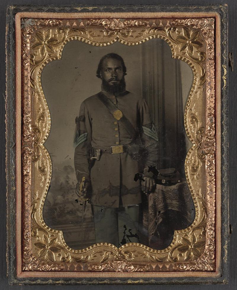 Unidentified African American soldier in Union infantry sergeant's uniform and black mourning ribbon with bayonet in front of painted backdrop, 1863 or 1865